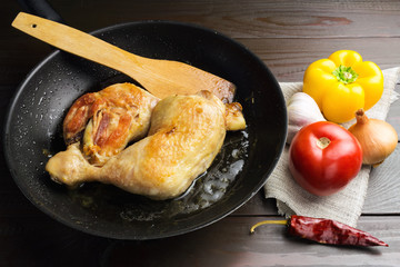 Two roasted chicken legs in black frying pan with vegetables and spices on dark wooden planks close-up view