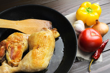Two roasted chicken legs in black frying pan with vegetables and spices on dark wooden planks close-up view