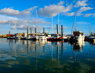 Fototapeta na wymiar Newhaven Marina, a row of 8 boats moored to a pontoon, the boats are white, the water is reflecting in the foreground, the sky blue with white fluffy clouds