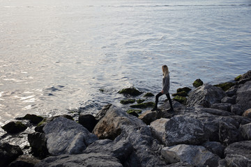 Young woman in warm clothes standing on stone on the beach. Girl enjoying au sunny autumn day and having fun outdoors. Lifestyle concept.