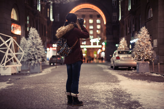Hipster girl walking through the night city street taking photos of buildings and Christmas trees