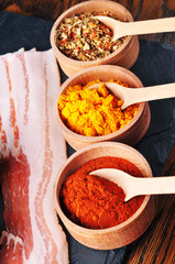 Bacon slices and wooden bowls with dry spices and spoons on the dark blue board on the wooden background
