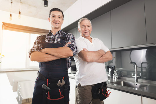 Two male plumbers posing in a modern kitchen. They have a special uniform, next to them a black box for the instrument.