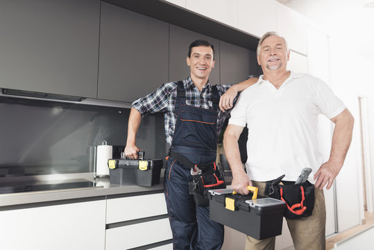 Two men of plumbers stand in the kitchen and pose with black toolboxes.