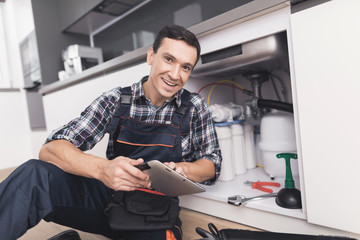 The plumber sits next to the kitchen sink on the floor and looks at the tablet for repair...