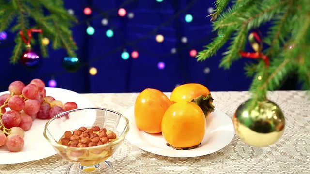 Plates with fruits and nuts with honey and are on the table with a decorated Christmas tree balls and flashing gerunds blurred bokeh. Photographed with dolly from right to left