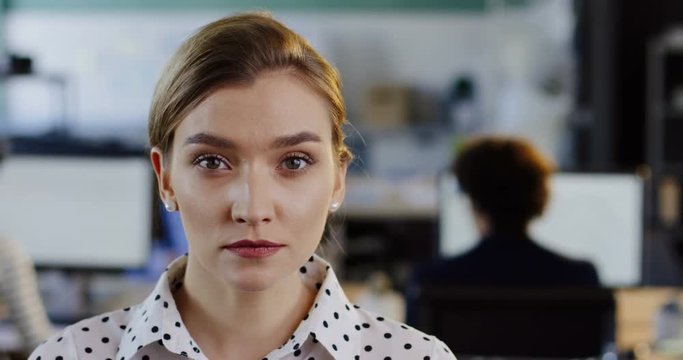 Close up of charming young woman turning her head to the camera and smiling sincerely. The blurred office with workers at computers background. Indoors. Portrait