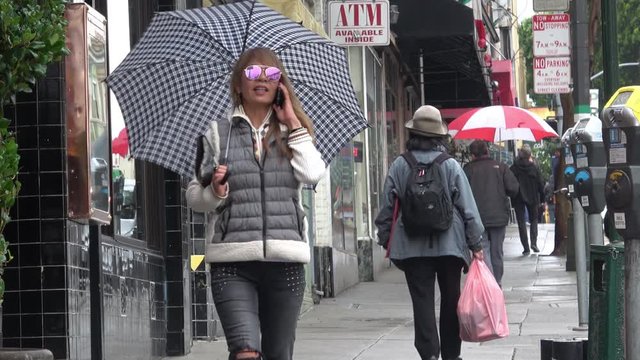 Rain in the City. Attractive Woman with an umbrella is walking down a busy street while talking on the phone on a rainy day. Footage from North Beach San Francisco.