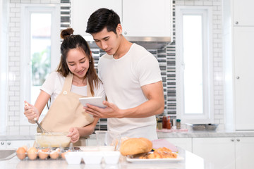 Couple cooking bakery in kitchen room, Young asian man and woman together making cake and bread with egg, looking menu from tablet in the flour happy relaxing in at home