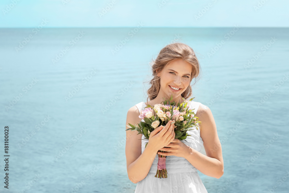 Wall mural Young smiling bride in white gown holding bouquet on seashore