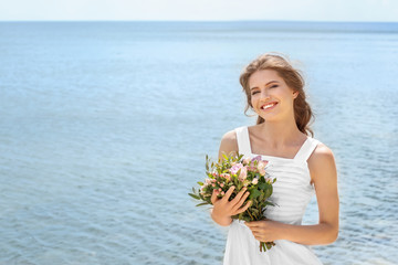 Young smiling bride in white gown holding bouquet on seashore