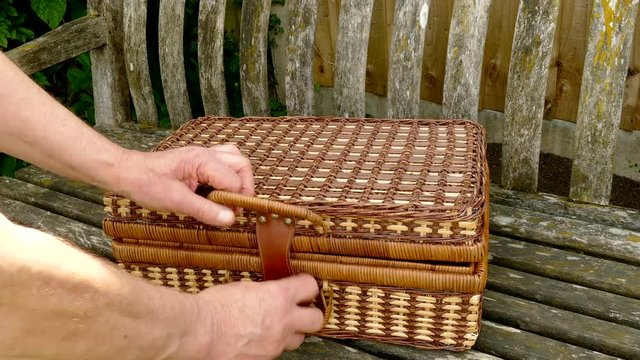 A man opening a wicker picnic hamper on an old wooden bench, and removing a cup and bottle of drink.
