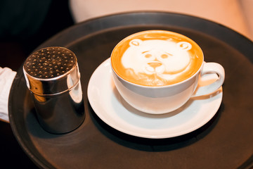 White cup of cappuccino with a picture of a bear's face, on a dark tray in the hand of an offspring close-up