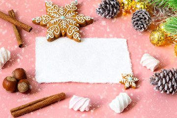 Obraz na płótnie Canvas Christmas greeting card over wooden light pink table with snow fir tree, snowflakes, nuts, marshmallow and cinamon. Flat lay slyle