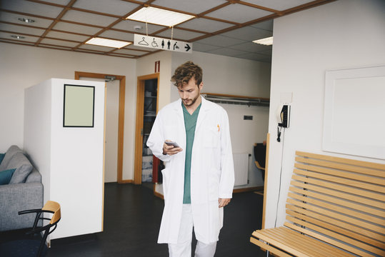 Young doctor using smart phone while walking in corridor at hospital