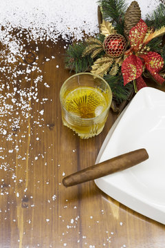 Christmas decoratoin on wooden background. Glass of whisky with ice, cigar in ashtrey decorated with snow.