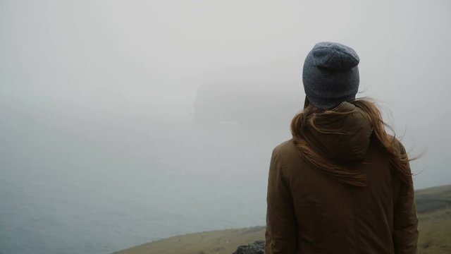 Back view of young woman in hat standing on the shore of the sea and thinking about something, exploring the Iceland.