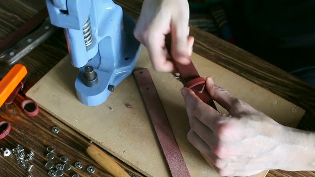 Master collects leather strap. On brown wooden table scattered with tools and accessories.