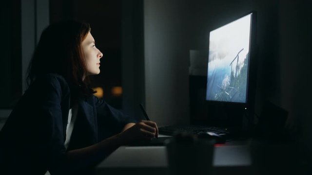 Concentrated young woman designer working in office at night using computer and graphics tablet to finish job