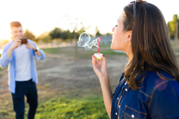 Fototapeta premium Handsome young man taking photo of his girlfriend making soap bubbles in the park.