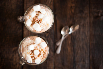 Two glasses with Hot chocolate garnished with whipped cream, marsmallow and cocoa powder.