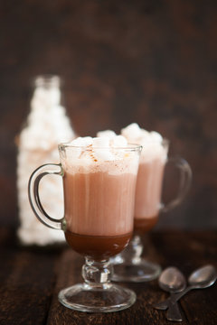 Two glasses with Hot chocolate garnished with whipped cream, marsmallow and cocoa powder.