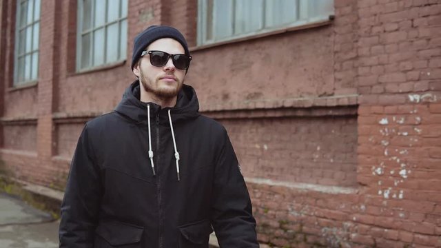Bearded hipster young man in black hat and sunglasses walking in urban background slow motion