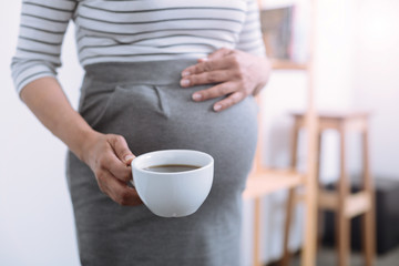 Big belly. Tender woman touching her baby abdomen and going to have break, drinking tea