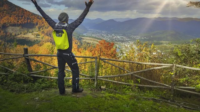 Young man standing on the edge of the cliff and raising his hands up against mountain range and bright colorful autumn forest. Traveler enjoying success and stunning view. Toning filter. Full HD