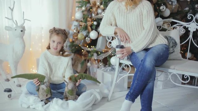 family Christmas photo session. Mom and daughter are sitting near the Christmas tree, in white knitted sweaters and jeans