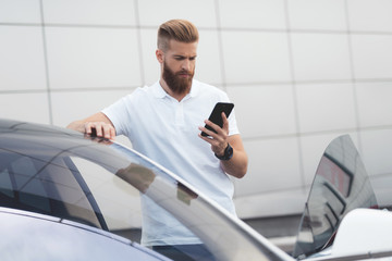 A young guy with a beard talking on the phone near his electric car.