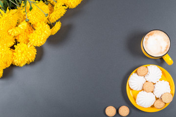 Food mock-up. Bright yellow plate with sweets (cookies and white marshmallows), big cup of cappuccino and bunch of chrysanthemums on gray background.Top view.