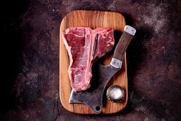 Fresh raw t-bones steak on an olive wooden board with sea salt and a kitchen ax. Top view.