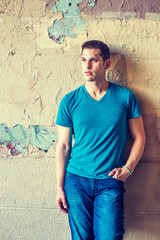 Fototapeta premium I miss You. A young American guy wearing blue V neck T shirt, jeans, stands on street by wall peeling off paints in hot summer, looking away, thinking, lost in thought.