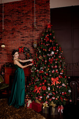 The smart brunette in an elegant green dress decorates a New Year's fir-tree. The girl is in the room with the fir-tree and a fireplace decorated by Christmas.