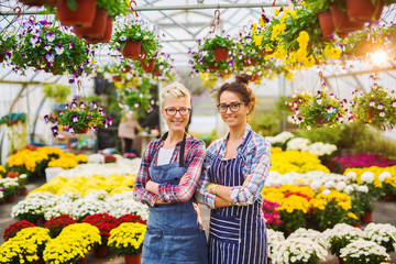 Two beautiful charming florist girls with eyeglasses and crossed hands standing in the sunny greenhouse full of flowers and looking at the camera.