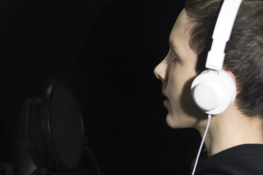 A cute teenager in white headphones records a voice in the dark room of the recording studio.
