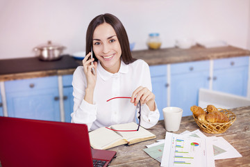 Beautiful young smiling business woman working at home behind laptop and talking on phone with client
