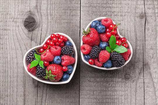 Berries overhead mix with leaves colorful closeup assorted in two heart shaped ceramic jars. Raspberry, blueberry, red currant, strawberry on rustic wooden table in studio.