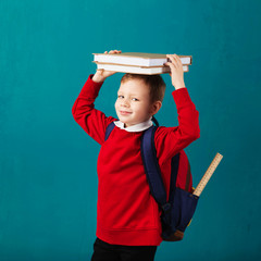 Fototapeta na wymiar Cheerful thoughtful little school boy in school uniform with backpack and big pile of books standing against blue wall. Looking at camera. School concept.