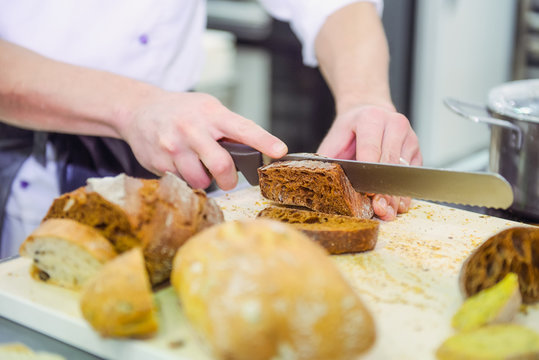 A male chef's hand cutting whole grain bread with knife on the table with various kinds of freshly baked breads. Selective focus, space for text