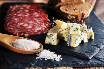 Roquefort slices with bread, sliced salami, salt on the spoon and peppercorns on the dark background