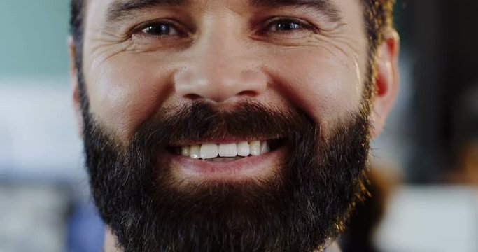 Close up of caucasias man with a beard looking and smiling sincerely straight into the camera. The blurred office with people at computers background. Inside. Portrait