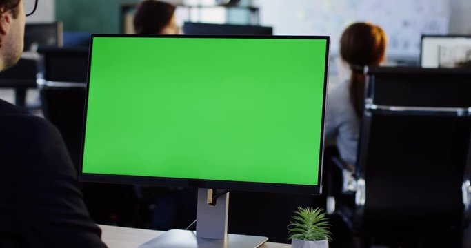 Male office employee working on the PC computer with green screen in the office room. View from behind. Chroma key. Indoors