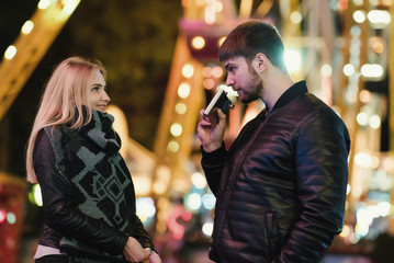 Young happy couple drinks whiskey alcohol from flask. Outdoors at night in an amusement park. Night city background 