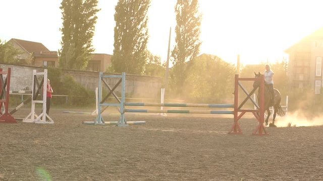 A young professional rider on a brown horse training jumping over obstacles. Slow motion. Sunset. Horse riding.