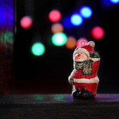 Dark christmas background with a Christmas tree toy a snowman  on a wooden board. Glare garlands of lights on a blurry side. Selective focus on the toy.