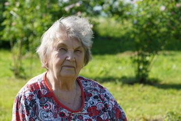 Portrait of old smiling woman dressed in t-shirt in the gargen in the background of growing trees in sunny summer day. Woman is charged energy of nature