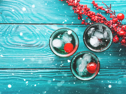 Christmas party green alcohol drinks with cherry. Festive aperitif shots and ornaments on wooden dark table. Holiday background with falling snow