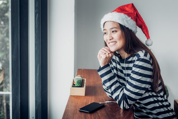 Asian teenage girl wear santa hat and smile face resting her chin on her hands near window in xmas party at cafe ,Celebrate New year and Christmas holiday concept.